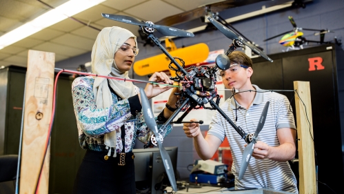 A male and female student work on a drone in a lab. The female is on the left, wearing black pants and a multi-colored top and a headscarf, the male to the right has brown hair and is wearing a gray polo shirt.