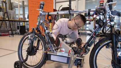 Male student wearing a light pink T-shirt works on a motorized bike in a lab setting.