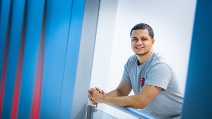 White male student leans against a railing and smiles. He has black hair and is wearing a gray Rutgers T-shirt.