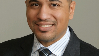 headshot of man with white and blue stripe collared shirt, blue, black, and grey, tie with a black jacket