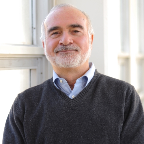 Head shot of balding male with white hair, moustache and beard, wearing a v neck pullover sweater with a collared button down shirt.