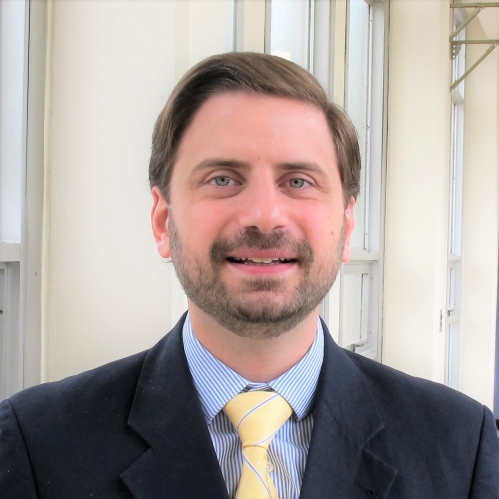Head shot of male with brown hair, moustache, and beard, wearing a dark suit jacket, with blue button down shirt, and yellow tie.