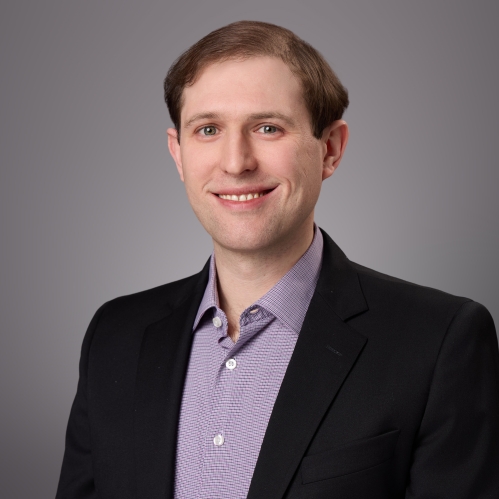 Headshot of male with short brown wavy hair wearing a black suit jacket with a patterned purple button down shirt.
