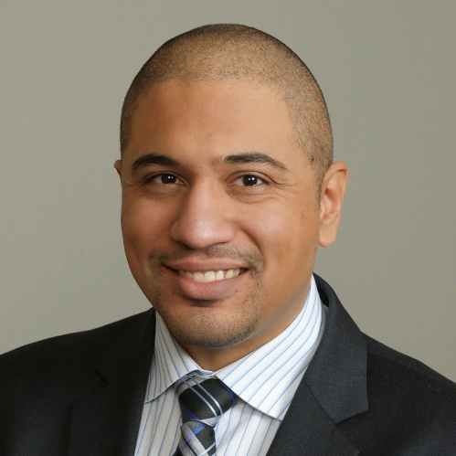 headshot of man with white and blue stripe collared shirt, blue, black, and grey, tie with a black jacket