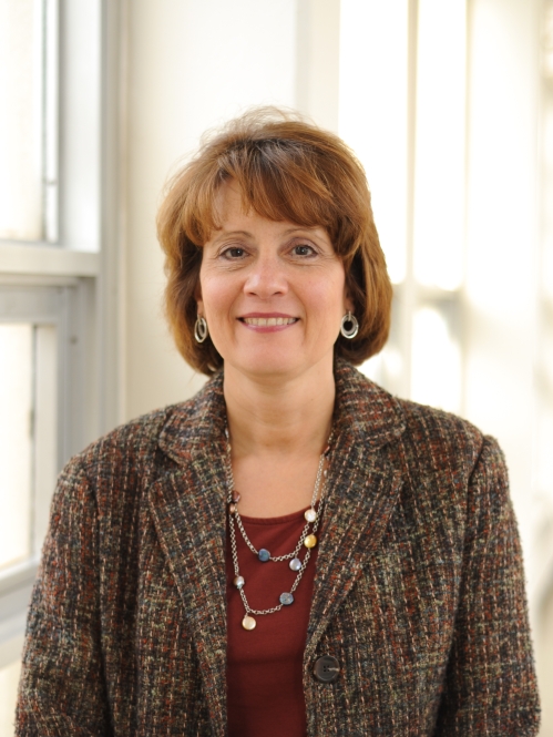 Head shot of woamn with brown hair, wearing a brown suit jacket, a maroon dress, necklace and earrings.