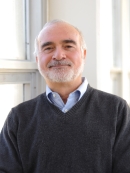 Head shot of balding male with white hair, moustache and beard, wearing a v neck pullover sweater with a collared button down shirt.