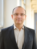 Head shot of balding male with eyeglasses, wearing a black suit jacket and a white button down shirt. 