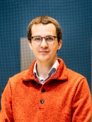 Head shot of male with eyeglasses wearing an orange collared pullover with buttons.