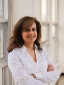 Head shot of female with her arms folded with long brown hair, wearing a white blouse 