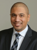 headshot of man with white and blue stripe collared shirt, blue, black, and grey, tie with a black jacket