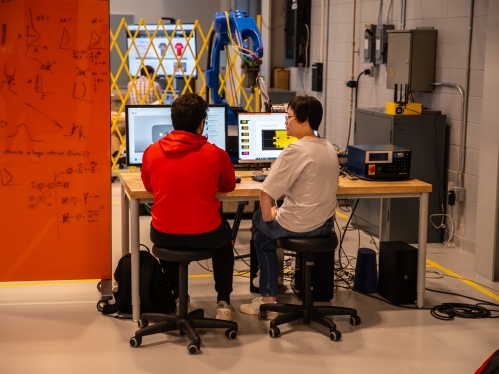 A female and male student sit at a desk with their backs to the camera. They are in a lab looking a desktop computer screens. To the left is an orange board with mathematical equations.