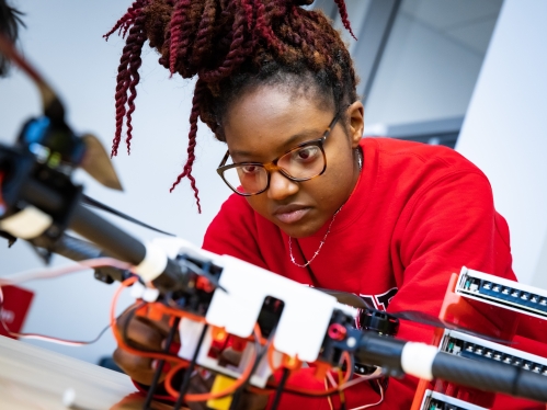 Black female engineering student works on a device. She wears a red sweatshirt, has glasses, and braids in a topknot bun.
