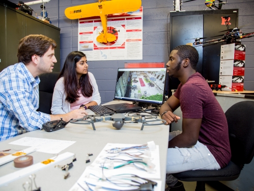 Two students, male and female, sit at a table with a professor discussing what they are looking at on a computer monitor.