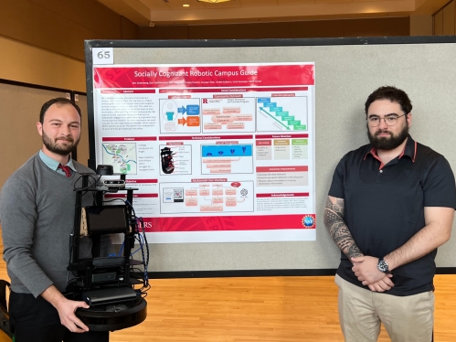 Two male students stand in front of a poster. The student on the left holds a robot while the other student stands with his arms. crossed.