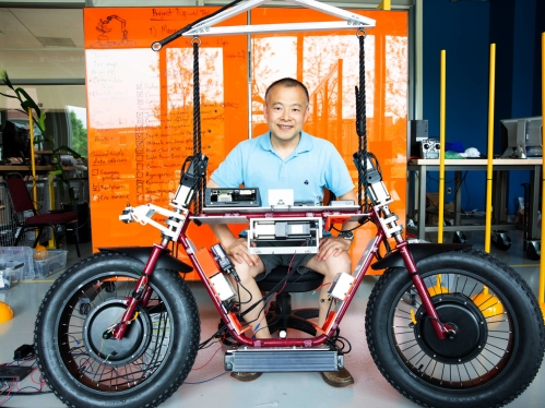 Male professor sitting behind motorcycle in robotics lab.
