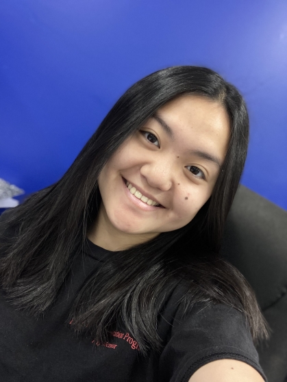 Young woman poses against a blue background. She has black hair and is wearing a black shirt.
