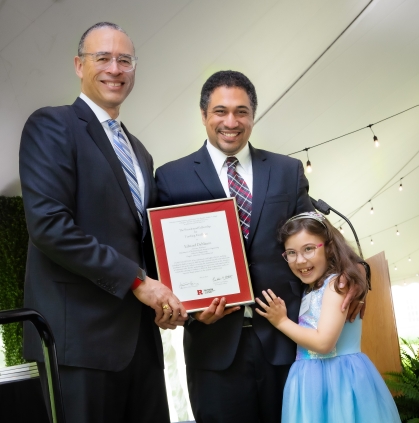 Rutgers University President, Jonathan Holloway on left presenting an award to a male faculty member alongside his young daughter.