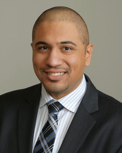 headshot of man with white and blue stripe collared shirt, blue, black, and grey, tie with a black jacket